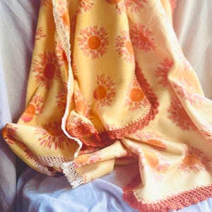 May include: A yellow fleece blanket with a repeating pattern of orange and pink sunburst designs. The blanket has a crocheted orange border and a white lace trim. The blanket is draped over a light blue surface.