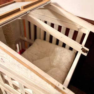 May include: White wooden cat house with a hinged roof and a soft white cushion inside. The roof has a mesh screen for ventilation.