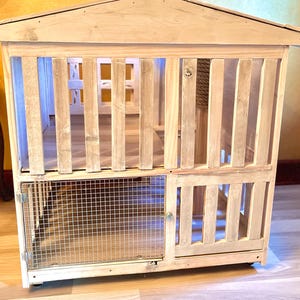 May include: A wooden pet house with a wire mesh door and a slatted wooden door. The house has a gabled roof and is on wheels.