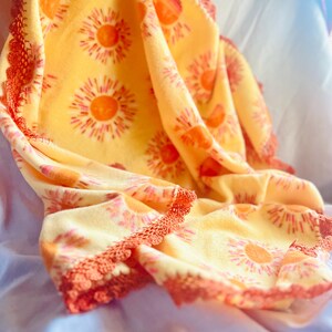 May include: A yellow blanket with a repeating pattern of orange suns. The blanket has a decorative orange lace trim around the edges. The blanket is folded and draped over a light blue surface.