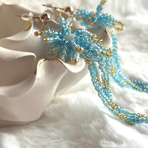 May include: Close-up of a pair of beaded earrings. The earrings feature a cluster of light blue beads with gold accents and dangling strands of beads. The earrings are displayed on a white surface.