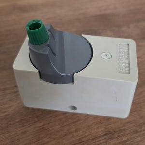 May include: A light gray, rectangular pencil sharpener with a dark gray top and a green nozzle. The text "EAGLE 7 TURQUOISE" is printed on the side. The sharpener is on a wooden surface.