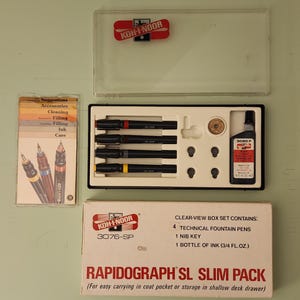 May include: A Koh-I-Noor Rapidograph SL Slim Pack set. The set includes four technical fountain pens, a nib key, and a bottle of ink (3/4 fl. oz.). The box also contains a clear-view box set. The set is designed for easy carrying.