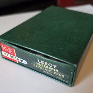 May include: A dark green box of Leroy Reservoir Lettering Pens. The box has a red and white label with the numbers "61 0050 05". The text "1/2 doz." is also visible on the box.