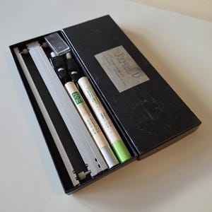 May include: A black rectangular box containing a drafting beam compass set. The set includes a silver metal beam, two black and silver compass heads, and two cylindrical tubes with labels reading K&E and Pleddl Beam Compass, Cincinnati, OH.