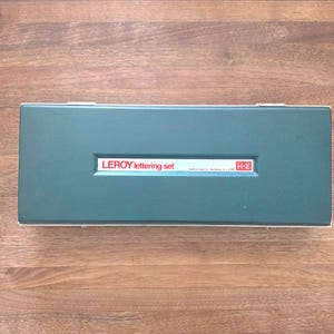 May include: A teal-colored LEROY lettering set case with a white rectangular label that reads "LEROY lettering set" in white text. The case is rectangular with a white border and a silver clasp. The case is closed and sits on a wooden surface.