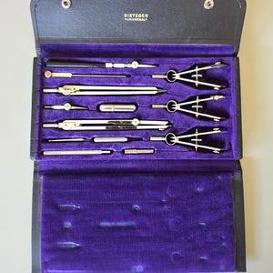 May include: A black leatherette case containing a set of silver metal drafting instruments, including compasses, dividers, and extension bars. The interior is lined with purple velvet, and the brand name Dietzgen Universal is printed in gold.
