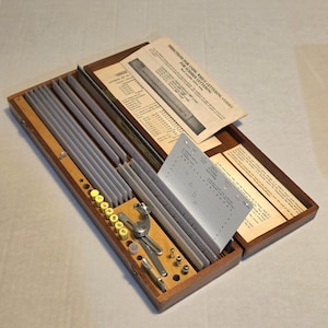 May include: A Wrico lettering set housed in a rectangular wooden box. The kit includes multiple metal lettering guides, a scriber tool, several yellow-capped ink pens, and a printed instruction manual titled Directions for Using Wrico Lettering Guides.