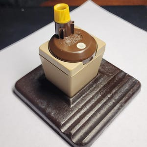 May include: A vintage brown and tan electrical outlet with a yellow top. The outlet is mounted on a dark brown, ridged base. The outlet has a small, circular sticker on the top.