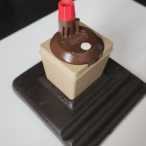 May include: A brown and tan electrical component with a red top, set on a dark brown, ridged base. The component has a circular brown top with a white dot and text. The base is square with rounded corners.