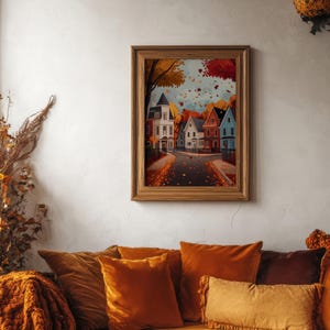 K&ouml;nnte beinhalten: Ein gerahmter Druck zeigt eine Stra&szlig;enszene mit bunten H&auml;usern und fallenden Herbstbl&auml;ttern. Das Kunstwerk befindet sich in einem Holzrahmen und h&auml;ngt &uuml;ber einem Sofa mit orangefarbenen Samtkissen und einer passenden Decke. Das Gesamtthema ist Herbstdekoration.