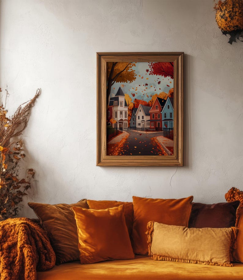 K&ouml;nnte beinhalten: Ein gerahmter Druck zeigt eine Stra&szlig;enszene mit bunten H&auml;usern und fallenden Herbstbl&auml;ttern. Das Kunstwerk befindet sich in einem Holzrahmen und h&auml;ngt &uuml;ber einem Sofa mit orangefarbenen Samtkissen und einer passenden Decke. Das Gesamtthema ist Herbstdekoration.