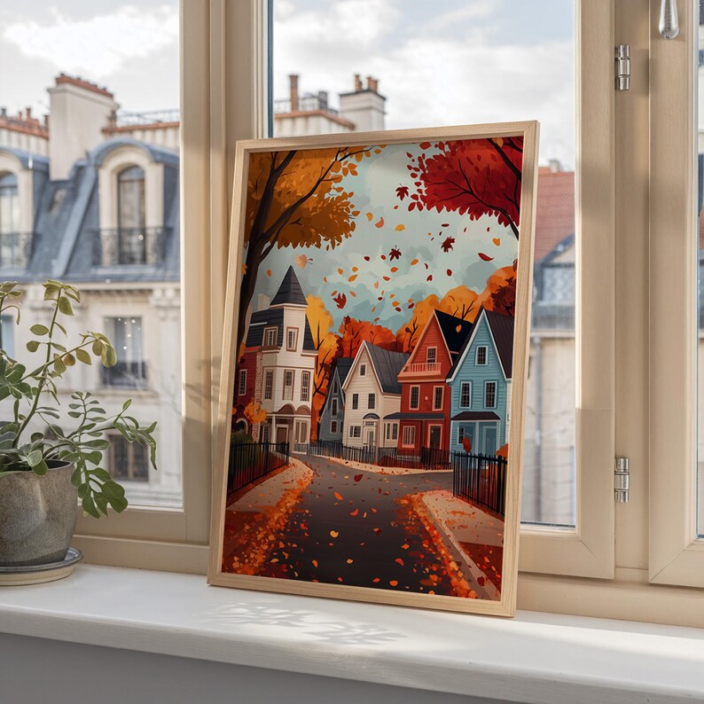 K&ouml;nnte beinhalten: Ein gerahmter Kunstdruck, der eine farbenfrohe Herbstszene zeigt. Das Kunstwerk zeigt eine Reihe von H&auml;usern in verschiedenen Farben, mit B&auml;umen in Orange- und Rott&ouml;nen. Gefallene Bl&auml;tter liegen auf der Stra&szlig;e und am Himmel verstreut.
