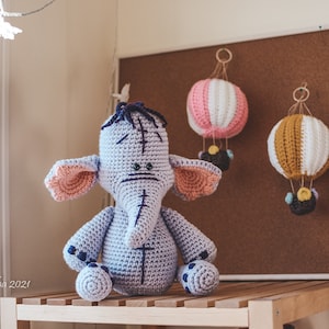 Puede incluir: Una muñeca de elefante de ganchillo hecha a mano con una expresión triste, sentada en un estante de madera. Tres globos aerostáticos de ganchillo de colores cuelgan en un corcho en el fondo.