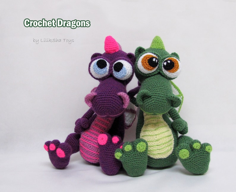 May include: Two crocheted dragon toys, one purple with pink accents and one green with yellow accents. Both dragons have large, googly eyes and are sitting on a white surface. The text "Crochet Dragons by Liliksha Toys" is visible above the dragons.