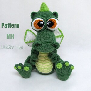 May include: Green crocheted dragon toy with large brown eyes and a white and green crocheted wing. The dragon is sitting with its legs crossed and has a green and yellow striped belly. The toy is on a white background.