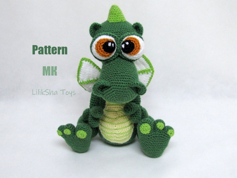 May include: Green crocheted dragon toy with large brown eyes and a white and green crocheted wing. The dragon is sitting with its legs crossed and has a green and yellow striped belly. The toy is on a white background.