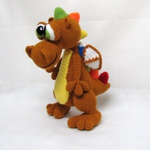 May include: A brown crocheted dragon toy with a green, orange, and yellow spiky crest, a white wing, and red tail. The dragon has a large, green eye and a friendly expression.