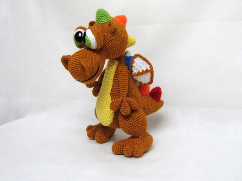 May include: A brown crocheted dragon toy with a green, orange, and yellow spiky crest, a white wing, and red tail. The dragon has a large, green eye and a friendly expression.