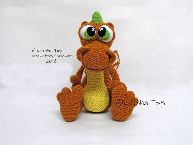 May include: A crocheted orange and yellow dragon toy with green horns and large white eyes. The dragon is sitting with its legs crossed and has a small white wing on its back. The text "@LilikSha Toys crochettoy.jimdo.com 2018" is visible on the image.