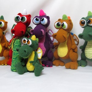 May include: A group of seven crocheted dragon toys in various colors, including green, red, purple, orange, and yellow. The dragons have large eyes and are standing on a white surface.