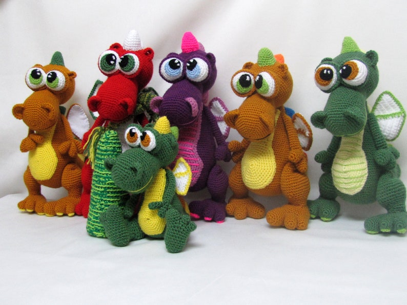 May include: A group of seven crocheted dragon toys in various colors, including green, red, purple, orange, and yellow. The dragons have large eyes and are standing on a white surface.