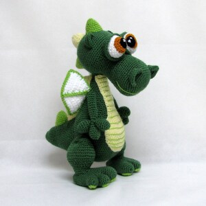 May include: Green crocheted dragon toy with large eyes and a white wing. The dragon has a light green stripe down its back.