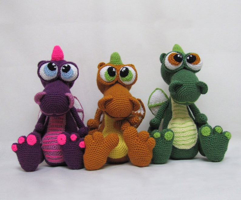 May include: Three crocheted plush dragons, one purple, one orange, and one green, with large eyes and a whimsical expression.