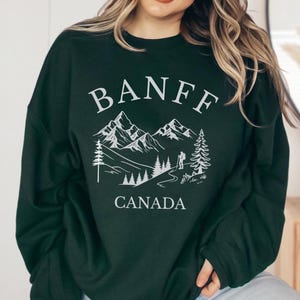 May include: A dark green crewneck sweatshirt featuring a white graphic print on the chest. The design displays the text BANFF in an arched font above a mountain range, pine trees, a hiking figure, and the word CANADA printed below the scene.