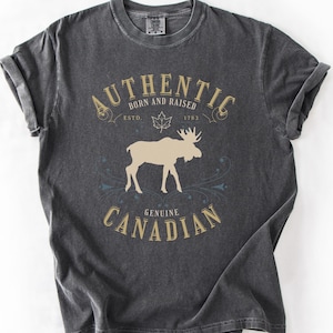 May include: A charcoal grey short-sleeve t-shirt featuring a beige moose silhouette graphic. The text reads AUTHENTIC BORN AND RAISED ESTD 1783 GENUINE CANADIAN, with a maple leaf icon above the moose and decorative flourishes surrounding the design.