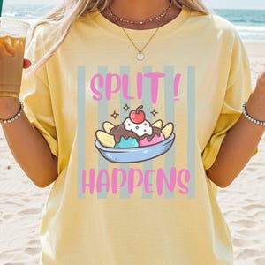 Può includere: Una t-shirt a maniche corte giallo pallido con una stampa grafica di una banana split in una coppetta con una ciliegia sopra. Il testo "SPLIT! HAPPENS" è stampato in rosa sopra e sotto l'illustrazione, su uno sfondo a righe verticali grigie.