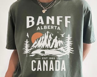 May include: An olive green short-sleeve t-shirt featuring a white graphic print of mountains, pine trees, a bear, and a sun. The text reads BANFF ALBERTA, EST 1885, and CANADA in a bold, sans-serif font centered on the chest of the garment.