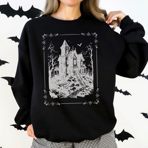 May include: A black long-sleeve sweatshirt featuring a white graphic print of a haunted house within a decorative rectangular frame. The house is surrounded by bare trees and a small bat, with a path leading to the front entrance of the building.