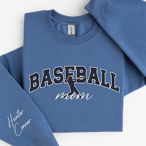 May include: A blue crew neck sweatshirt featuring the word "BASEBALL" in a navy blue block font with a white outline. A silhouette of a baseball player is in the centre. The word "mom" is written in cursive below. The left sleeve has the names "Hunter Connor" in cursive.