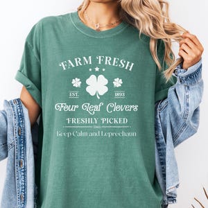 May include: Green t-shirt featuring the text "FARM FRESH" and "Four Leaf Clovers FRESHLY PICKED" in white. The design includes a four-leaf clover and the phrase "Keep Calm and Leprechaun". The shirt has rolled-up sleeves.