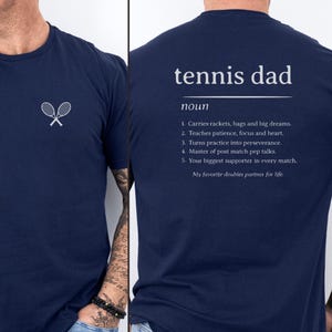 May include: Navy blue t-shirt with a small white tennis racket logo on the front. The back of the shirt features the text "tennis dad" above a definition, including phrases like "carries rackets" and "biggest supporter."
