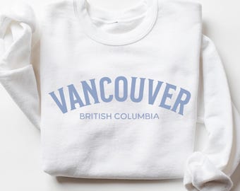 May include: A white crewneck sweatshirt featuring the text VANCOUVER in a large, arched, light blue font, with the words BRITISH COLUMBIA printed in a smaller, straight, light blue font directly underneath the city name.