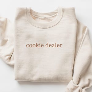 May include: A cream-coloured crewneck sweatshirt featuring the text "cookie dealer" embroidered in a brown, serif font across the centre of the chest. The garment has long sleeves and a ribbed collar, cuffs, and hem, presented in a folded layout.
