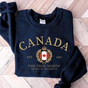 May include: A navy blue crewneck sweatshirt featuring gold text that reads CANADA, EST 1867, and THE TRUE NORTH. Below the text is a circular Canadian flag emblem surrounded by a gold laurel wreath, a small crown, and the coordinates 56.1304°N, 106.3468°W.