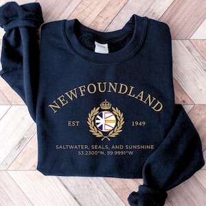May include: A navy blue crewneck sweatshirt featuring gold text that reads NEWFOUNDLAND, EST 1949, and SALTWATER, SEALS, AND SUNSHINE 53.2300°N, 59.9991°W. A central gold laurel wreath surrounds a small circular flag icon with a crown above it.