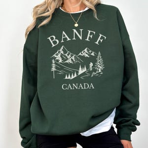 May include: A dark forest green crewneck sweatshirt featuring a white graphic print on the chest. The design displays the text BANFF in an arched font above a mountain landscape illustration with pine trees and a hiker, with CANADA below.