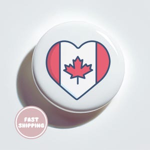 May include: A white button badge with a heart-shaped design of the Canadian flag. The flag features a red maple leaf on a white background, framed by red bars. The badge has a blue outline.