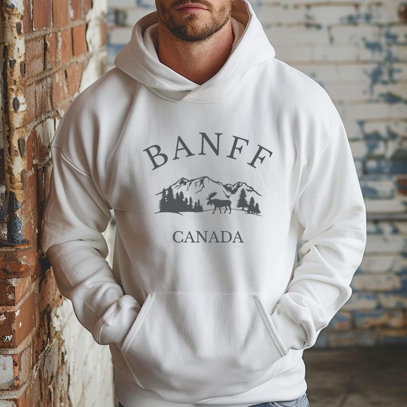 A man wearing a white hoodie with a dark grey graphic that reads BANFF CANADA above an illustration of mountains, trees, and a moose.