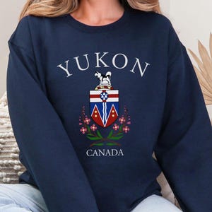 Könnte beinhalten: Marineblaues Sweatshirt mit dem Wort "YUKON" in Weiß, das sich oben in einem Bogen befindet. Darunter befindet sich ein Wappen mit einem Hund und das Wort "CANADA" unten. Das Wappen hat rote, weiße und blaue Elemente mit rosa Blumen.