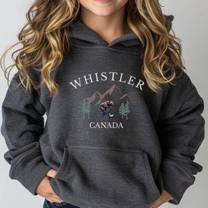 May include: A dark charcoal gray hooded sweatshirt featuring a graphic print with the text "WHISTLER" above and "CANADA" below a mountain range, green pine trees, and a brown bear riding a bicycle in the center, with a front kangaroo pocket.