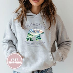 Peut inclure: Sweat-shirt à capuche gris avec un motif graphique représentant une silhouette d'ours avec un paysage de montagne à l'intérieur. Le texte "Canadian Girl" est au-dessus de l'ours et "Now and Forever" est en dessous de l'ours. Le texte "Since 1976" est entre l'ours et le texte "Now and Forever". Le texte "True North Strong and Free" est en dessous du texte "Now and Forever".