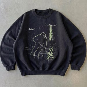 May include: A black crewneck sweatshirt featuring a graphic print of a large, hairy creature playing ice hockey on a rink. Above the creature, a flying saucer emits a bright green beam of light that illuminates the ice and the hockey stick.
