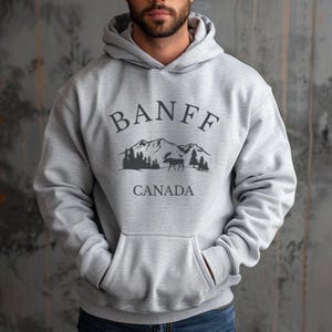 A man wearing a grey hoodie with a  black graphic that reads BANFF CANADA above an illustration of mountains, trees, and a moose.