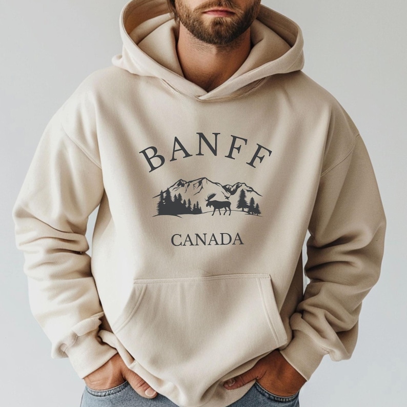 A man wearing a grey hoodie with a  black graphic that reads BANFF CANADA above an illustration of mountains, trees, and a moose.
