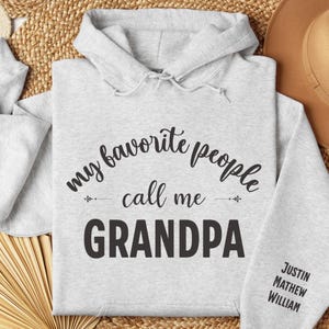 May include: Pale grey hoodie featuring the words "my favourite people call me GRANDPA" in black script. The sleeve displays the names "JUSTIN, MATHEW, WILLIAM" in black. The hoodie has a hood with drawstrings.